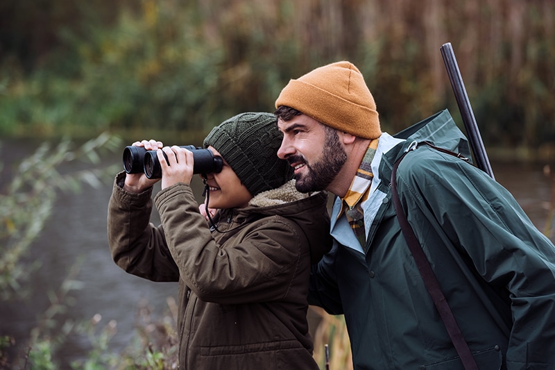 Son looking through binoculars, father standing with a gun